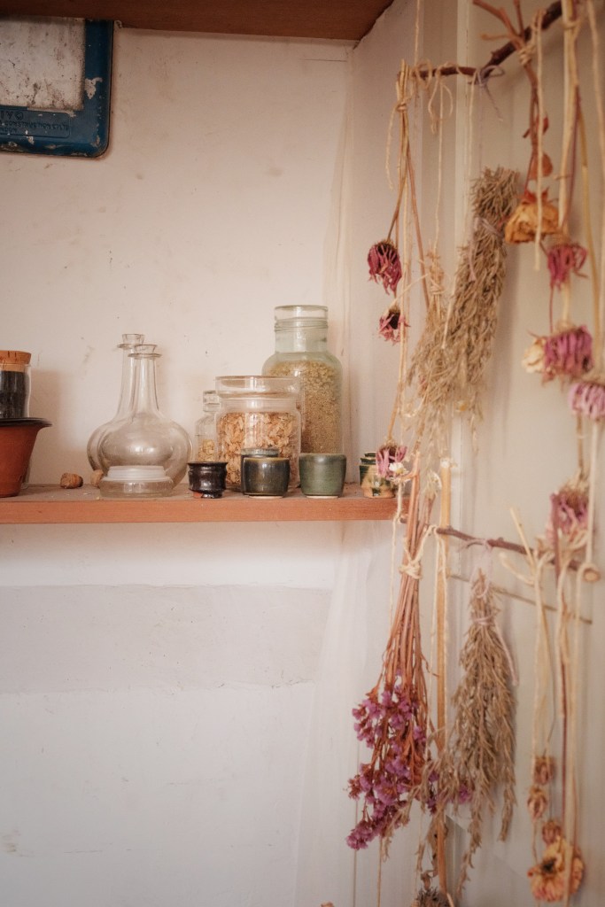 A wooden shelf displaying various jars and glass containers filled with dried herbs and spices, alongside a small terracotta pot, and adorned with dried flowers hanging on strings.